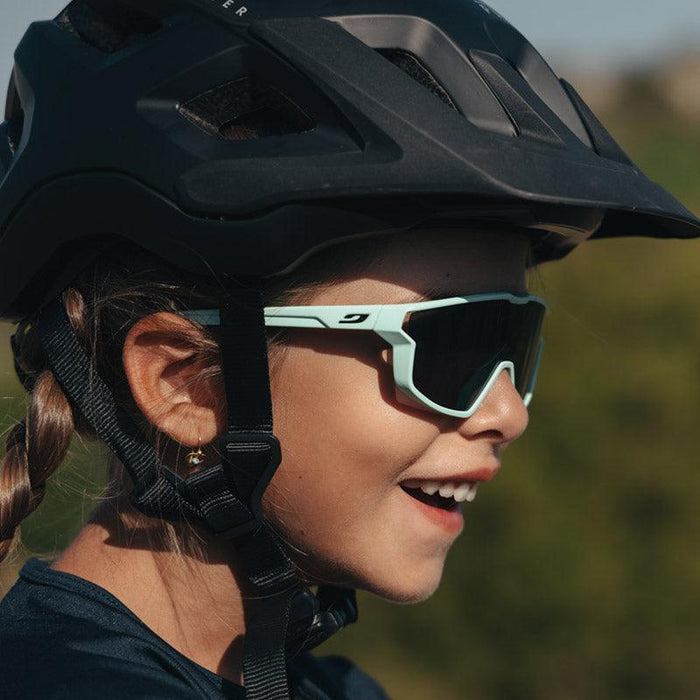 A young child wearing a black cycling helmet and Julbo-usa Fury Mini Kids sunglasses smiles outdoors with a blurred green background.