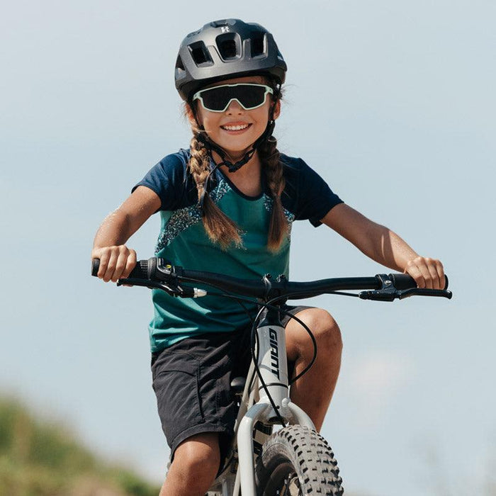 A young girl wearing a bike helmet, Julbo-usa Fury Mini Kids sunglasses, and a sports outfit rides her bicycle on a sunny outdoor pump track.