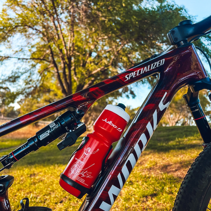 Close-up of a maroon Specialized S-Works bike frame with a red Julbo Purist Water Bottle by Julbo Eyewear, featuring a Moflo 2.0 cap, set outdoors on grass with trees in the background. #color_haley-batten-olympian-26oz-red