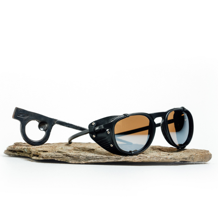 A pair of black Julbo Eyewear Limited Edition Millenium sunglasses with side shields and round arm cutouts, polarized Category 3 lenses, and a hand-forged piton bottle opener, displayed on a flat stone surface.