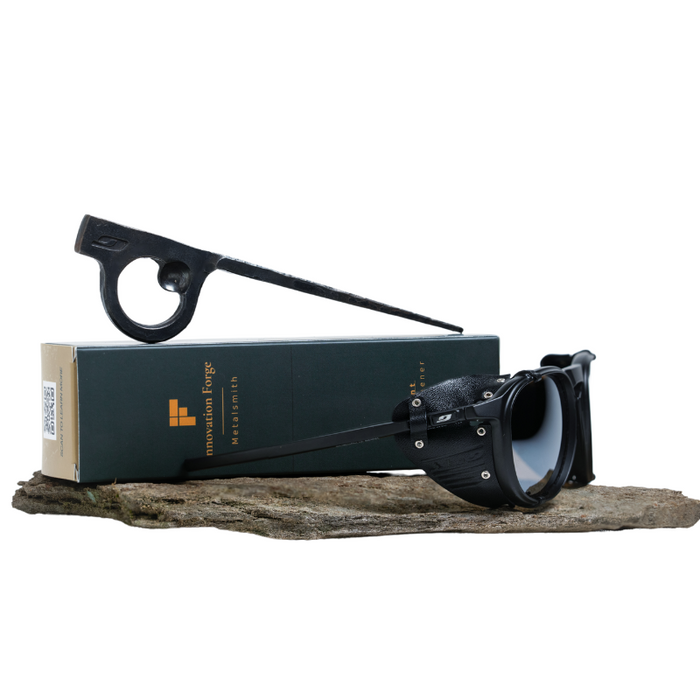 A pair of Julbo Eyewear's black Limited Edition Millenium goggles with polarized Category 3 lenses and a hand-forged Piton Bottle Opener rest on a green Mountain Forge box on stone, echoing mountaineering heritage.