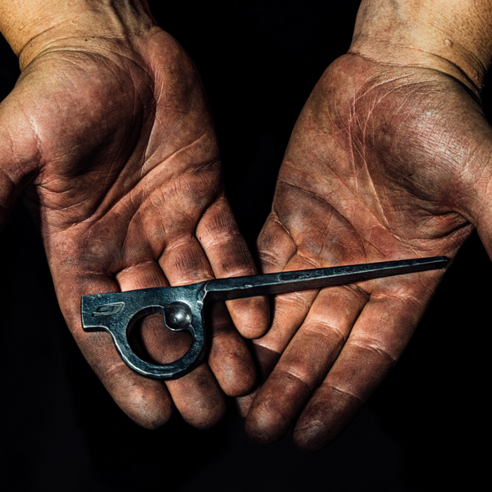 A pair of dirty, calloused hands holds Julbo Eyewear's Limited Edition Millenium with Hand Forged Piton Bottle Opener against a dark background, evoking the brand’s mountaineering heritage.