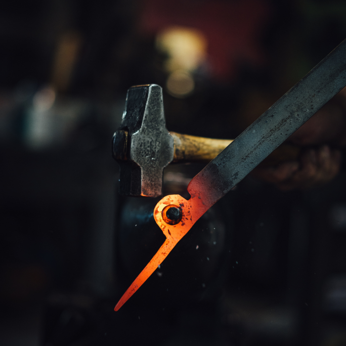 In a blacksmithing workshop, someone wearing Julbo Eyewear’s Limited Edition Millenium glasses forges metal with a hammer and anvil, drawing on mountaineering heritage—just like the included Hand Forged Piton Bottle Opener.