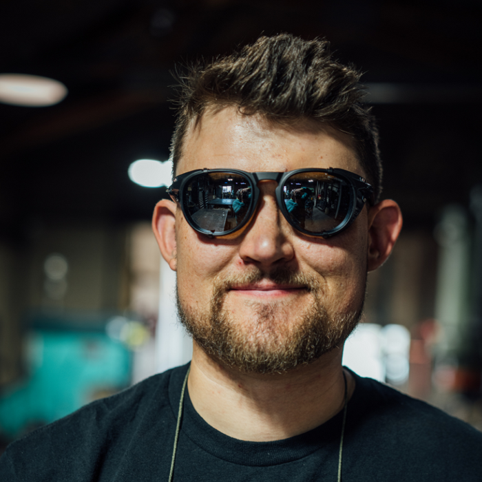 A bearded man with short hair wears Julbo Eyewear’s Limited Edition Millenium sunglasses with Hand Forged Piton Bottle Opener and a black shirt, standing indoors with blurred industrial equipment in the background.