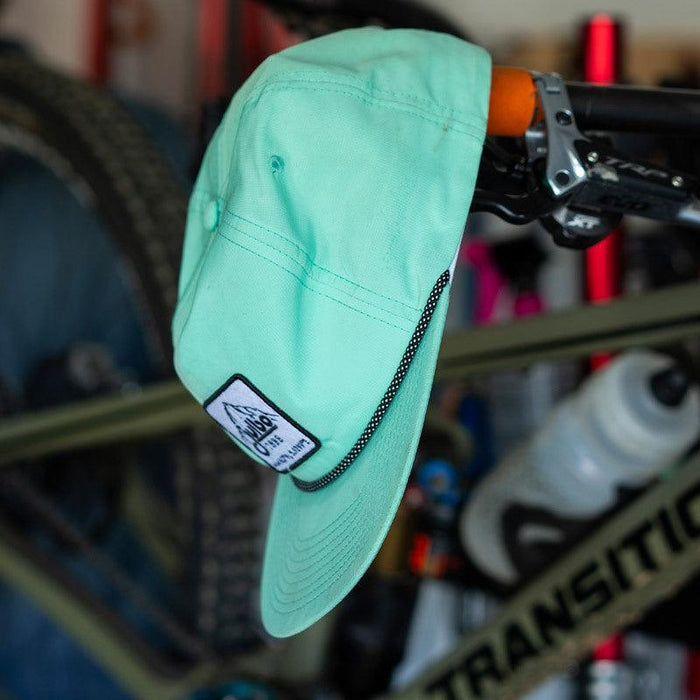 A mint green Julbo Eyewear Né dans les Alpes Unstructured Hat with a logo patch hangs on a bike handlebar, with bicycle parts and a water bottle in the background. #color_né-dans-les-alpes-unstructured-hat-bluebird