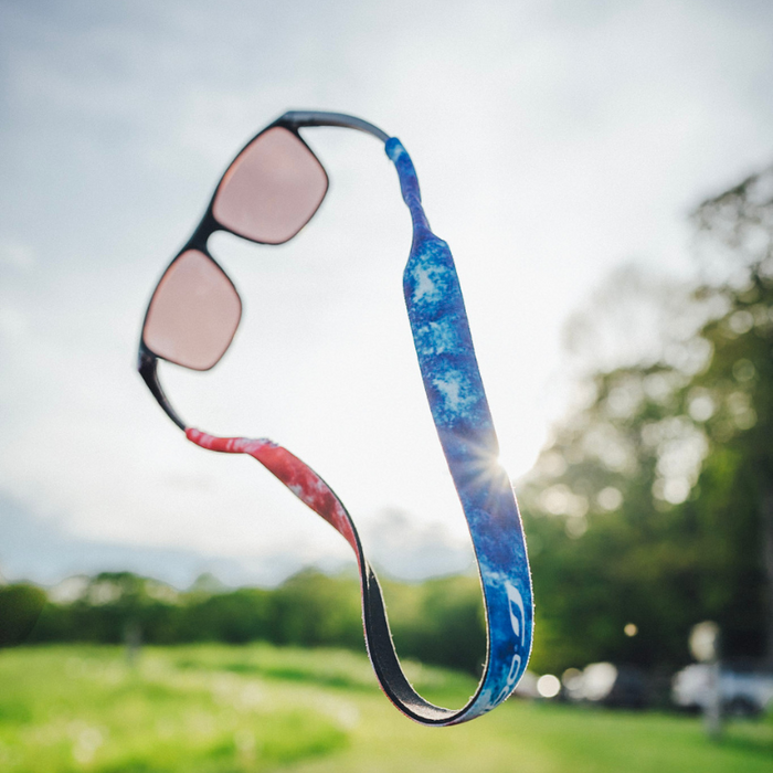 A Julbo Eyewear Neoprene Sunglass Strap in blue and red holds a pair of sunglasses that appear to float outdoors, surrounded by trees and sunlight. #color_julbo-neoprene-sunglass-strap-star-spangled